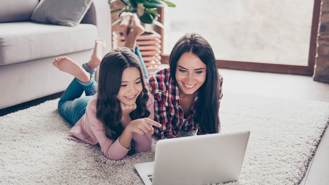 Mother and her little curious daughter are lying on a floor at home and using a laptop