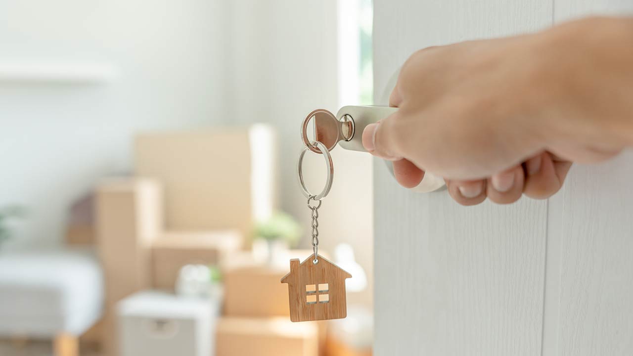 A key inserted into a door of the new house with a hand opening it, inside the room are cardboard boxes and furniture