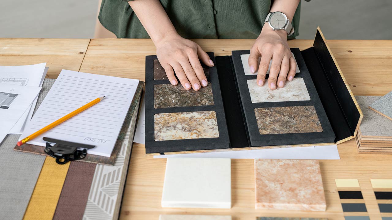 Hands of designer on samples of marble tile surrounded by wallpaper patterns during work over new project or order of client